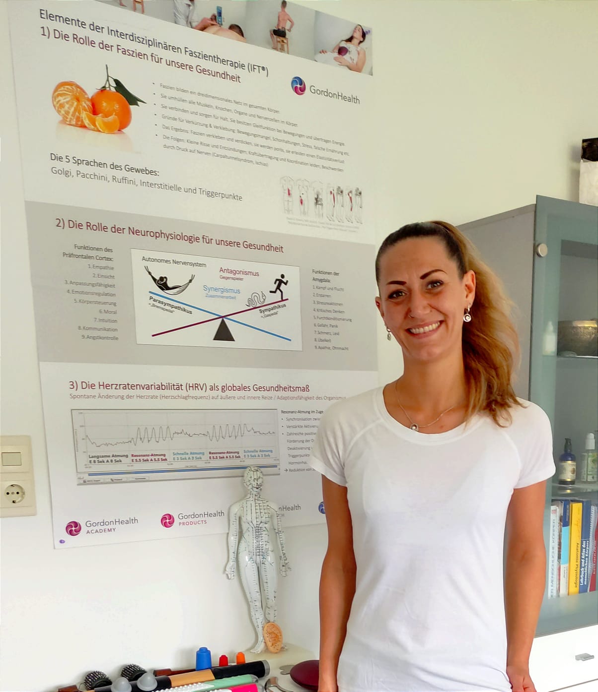 Maya in ihrer Praxis vor einem Poster zur Interdisziplinären Faszientherapie (IFT®)
